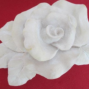 May include: A detailed, off-white rose sculpture with multiple petals and leaves. The sculpture is set against a red background, highlighting the intricate details of the floral design. The rose is a classic decorative element.
