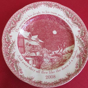 May include: A round, white ceramic plate with a red winter scene and text. The plate features a house, sleigh, and moon illustration. The rim has a holly and berry design. The text reads "He sprang to his sleigh..." and "2008".