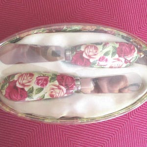 May include: A set of two metal spreaders with floral patterned handles featuring red and pink roses on a cream background. The spreaders are housed in an oval-shaped clear plastic container lined with white fabric, displaying a matching rose pattern.