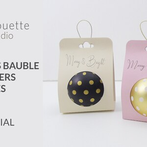 Op de afbeelding: Twee kerstbalhouders, een beige en een roze, met uitgesneden cirkels om kerstballen te houden. De beige houder heeft de tekst "Merry & Bright!" en de roze houder heeft de tekst "Merry Christmas!"