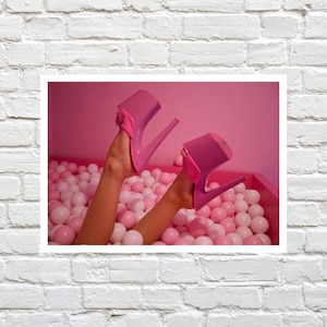 Könnte beinhalten: Ein Paar pinkfarbene Plateau-High Heels werden in die Luft gehalten, wobei die Füße einer Person sichtbar sind. Die Absätze befinden sich in einer rosa Bällchengrube mit rosa und weißen Bällen. Der Hintergrund ist einfarbig rosa.
