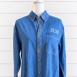 May include: A long-sleeved blue denim button-down shirt featuring a classic collar, a single chest pocket on the left side, and metallic buttons. The letters JLM are embroidered in white thread above the chest pocket on the front of the shirt.