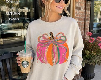 May include: A beige long-sleeve crewneck sweatshirt featuring a central graphic of a pumpkin decorated with a large, multi-colored bow. The pumpkin design incorporates vibrant brushstrokes of orange, pink, and green, accented with gold glitter details.