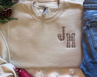 May include: A beige crewneck sweatshirt featuring embroidered letters JM in a red, white, and green plaid pattern on the left chest. The sweatshirt is paired with blue denim jeans, a small green pine branch with a pinecone, and a red bell ornament.
