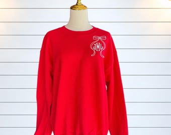 Puede incluir: Una sudadera roja de cuello redondo con un diseño bordado en blanco en la parte superior izquierda del pecho. El diseño consiste en el contorno de un lazo decorativo con las letras del monograma JML centradas en una fuente serif.