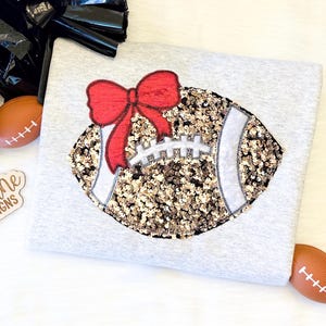 May include: A light gray sweatshirt featuring a football design with a red bow. The football is embellished with gold and silver sequins. A black pom-pom and two miniature footballs are also visible.