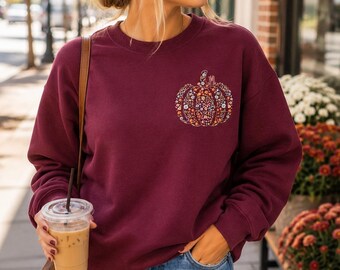 May include: A maroon long-sleeve crewneck sweatshirt featuring a pumpkin graphic on the left chest. The pumpkin design is filled with a colorful floral pattern and includes the monogram letters JML at the top. The sweatshirt has a ribbed neckline.