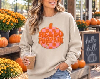 May include: A beige crewneck sweatshirt featuring a large, circular graphic on the chest. The graphic displays a pink and orange checkered pumpkin pattern with the text "HELLO PUMPKIN" in bold, orange, retro-style lettering across the center.