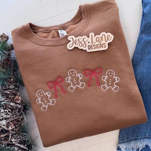 May include: A brown sweatshirt with embroidered gingerbread men and red bows. The sweatshirt has the text "Jess Lane Designs" on a wooden tag. The design is on the front of the sweatshirt. Denim jeans are visible in the background.