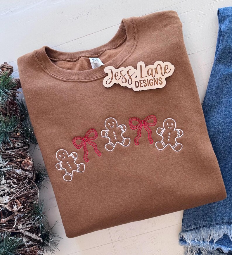 May include: A brown sweatshirt with embroidered gingerbread men and red bows. The sweatshirt has the text "Jess Lane Designs" on a wooden tag. The design is on the front of the sweatshirt. Denim jeans are visible in the background.