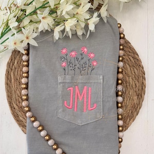 May include: A gray t-shirt with a pocket embroidered with pink flowers and the initials "JML". The shirt is displayed on a woven mat with a beaded garland and white flowers.