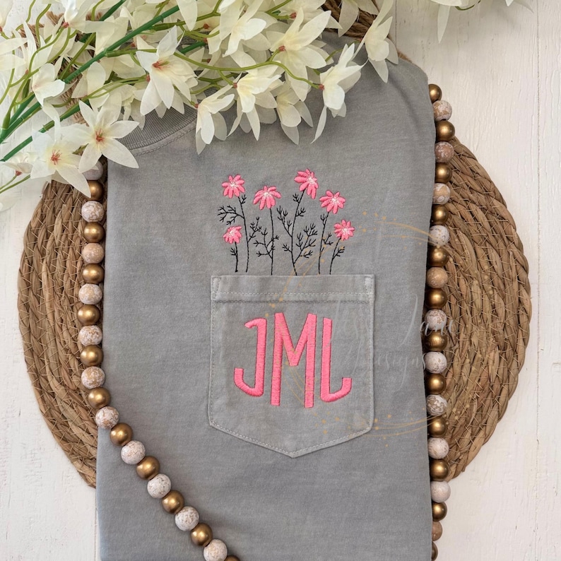 May include: A gray t-shirt with a pocket embroidered with pink flowers and the initials "JML". The shirt is displayed on a woven mat with a beaded garland and white flowers.