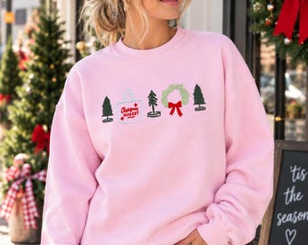 Puede incluir: Una sudadera de cuello redondo rosa claro con un diseño bordado de tres pinos verdes, una bolsa de tela blanca con el texto rojo Christmas Market y una corona verde con un lazo rojo centrado en la zona del pecho de la prenda.