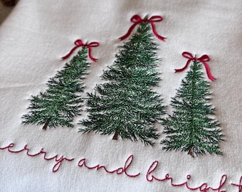 May include: A white sweatshirt featuring an embroidered design of three green pine trees, each topped with a small red ribbon bow. Below the trees, the text "merry and bright" is embroidered in red script. A wooden tag reads "Jess Lane DESIGNS".