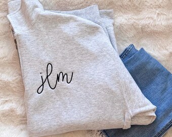 May include: A light gray quarter-zip sweatshirt featuring a high collar and a silver-toned zipper. The front of the garment is embroidered with the lowercase black script letters jlm. A pair of blue denim jeans is folded underneath the sweatshirt.