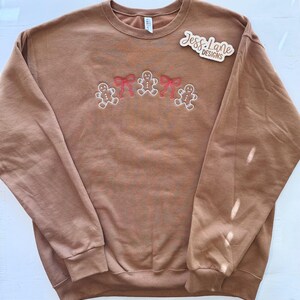 May include: A brown sweatshirt with embroidered gingerbread men and red bows. The sweatshirt has a crew neck and long sleeves. The text "Jess Lane Designs" is on a wooden sticker on the upper right side of the sweatshirt.