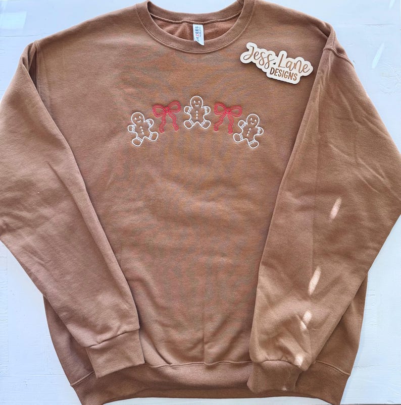 May include: A brown sweatshirt with embroidered gingerbread men and red bows. The sweatshirt has a crew neck and long sleeves. The text "Jess Lane Designs" is on a wooden sticker on the upper right side of the sweatshirt.