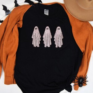 May include: Black raglan shirt with three embroidered pink ghosts. The shirt has orange sleeves and is part of a Halloween theme. A brown hat and a plastic spider are also visible.