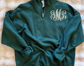 May include: A dark green quarter-zip sweatshirt featuring a high collar and long sleeves with ribbed cuffs. The left chest area displays a large, white, decorative monogram with the letters SML in a flowing, cursive script font style.