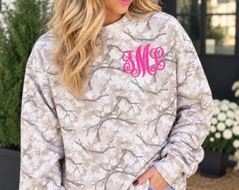 May include: A long-sleeve crewneck sweatshirt featuring a light beige and gray tree branch camouflage pattern. The garment displays a bright pink embroidered monogram on the upper left chest area, paired with light blue denim shorts.