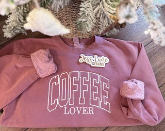 May include: A dusty rose crewneck sweatshirt featuring the white text COFFEE LOVER in a collegiate font. The interior of the garment is visible at the cuffed sleeves, showing a soft, fuzzy fleece lining. A small white logo tag reads Jess Lane DESIGNS.