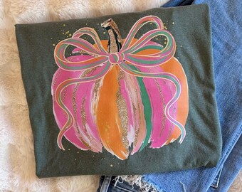 May include: An olive green t-shirt featuring a graphic of a pumpkin painted in vertical stripes of bright pink, orange, and green with gold glitter accents. A large, multi-colored striped bow is tied around the stem of the pumpkin.