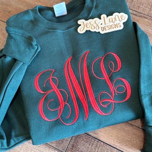 May include: A dark green crewneck sweatshirt featuring a large, red embroidered monogram with the letters M and L in a cursive script font. A white tag is visible at the neckline, and a wooden sign reads Jess Lane Designs.