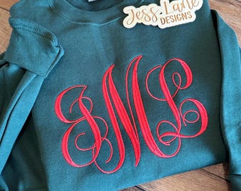 May include: A dark green crewneck sweatshirt featuring a large, red embroidered monogram with the letters M and L in a cursive script font. A white tag is visible at the neckline, and a wooden sign reads Jess Lane Designs.