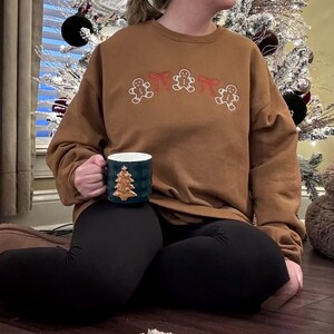May include: A brown sweatshirt with embroidered gingerbread men and red bows. A dark green mug with a gold Christmas tree design is held in the hand. The person is wearing black leggings.