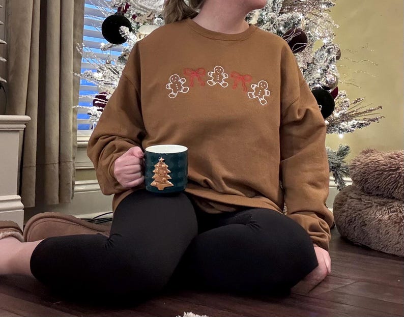 May include: A brown sweatshirt with embroidered gingerbread men and red bows. A dark green mug with a gold Christmas tree design is held in the hand. The person is wearing black leggings.