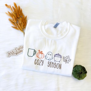 May include: White sweatshirt with embroidered fall-themed designs, including a mug, pumpkin, ghost, cauldron, and leaf. The phrase "COZY SEASON" is embroidered below the images. A sprig of dried flowers and a small green pumpkin are also present.