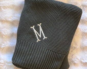 May include: A dark gray knitted blanket featuring a textured garter stitch pattern and a wide ribbed border. The center of the blanket is embroidered with a white capital letter M in a serif font, creating a clear contrast against the dark fabric.