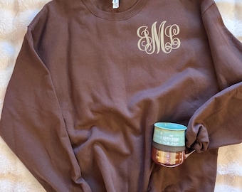 May include: A solid brown crewneck sweatshirt featuring a light tan monogram with the letters SML in a decorative script font on the upper left chest. A two-toned mug with a teal top and a dark red bottom is placed over the lower right side.