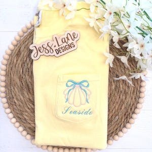 May include: A yellow t-shirt with a pocket featuring a seashell design and the word "Seaside" embroidered in blue. The shirt has a light blue bow and pink seashell design. The wooden sign says "Jess Lane Designs".