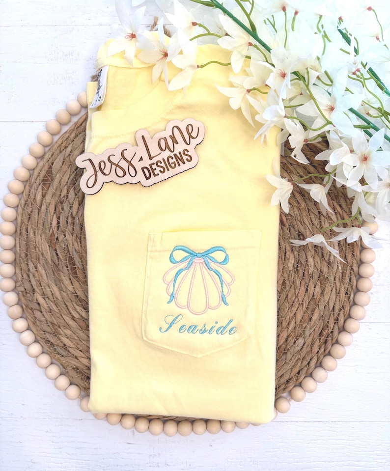May include: A yellow t-shirt with a pocket featuring a seashell design and the word "Seaside" embroidered in blue. The shirt has a light blue bow and pink seashell design. The wooden sign says "Jess Lane Designs".