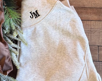 May include: A light heather gray sweatshirt featuring a high collar and long sleeves. The upper left chest area is embroidered with the black capital letters JM in a serif font. The garment is folded neatly, showcasing the textured fabric.