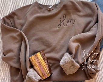 May include: A brown crewneck sweatshirt featuring the embroidered lowercase letters jlm on the upper left chest. The sweatshirt has long sleeves with folded cuffs revealing a light tan fleece lining and is paired with a copper-colored tumbler.