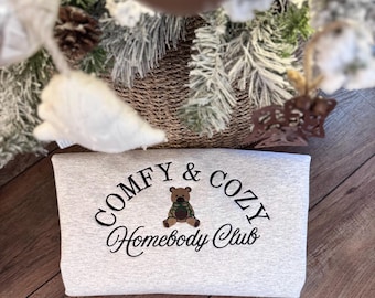 May include: A light gray heather sweatshirt featuring black embroidered text that reads COMFY & COZY Homebody Club. Centered between the words is a small brown teddy bear wearing a green and black plaid shirt. The design is stitched on the front.