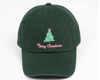 May include: A dark green baseball cap featuring an embroidered light green Christmas tree decorated with small pink bows. Below the tree, the text Merry Christmas is embroidered in pink script lettering on the front panel.