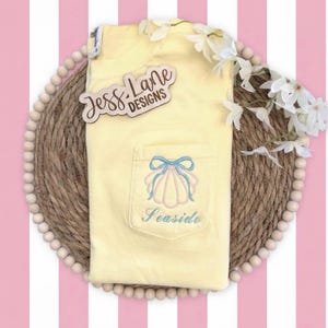 May include: A light yellow pocket t-shirt with a seashell design embroidered on the pocket. The design includes a light blue bow and the word "Seaside". The shirt is on a woven mat with white flowers.