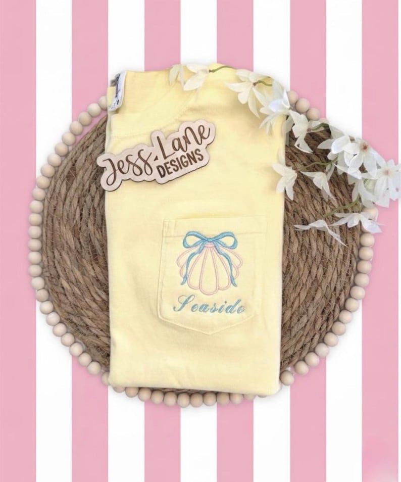 May include: A light yellow pocket t-shirt with a seashell design embroidered on the pocket. The design includes a light blue bow and the word "Seaside". The shirt is on a woven mat with white flowers.