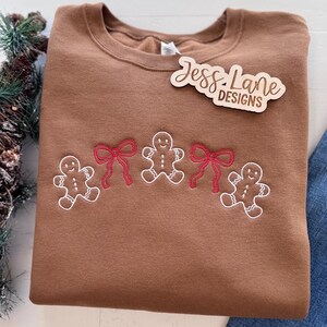 May include: A brown crewneck sweatshirt with embroidered gingerbread men and red bows. The sweatshirt has the text "Jess Lane Designs" on a wooden tag. The design is a festive holiday theme.