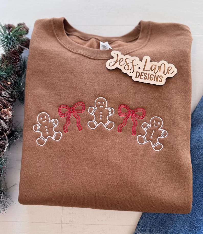 May include: A brown crewneck sweatshirt with embroidered gingerbread men and red bows. The sweatshirt has the text "Jess Lane Designs" on a wooden tag. The design is a festive holiday theme.