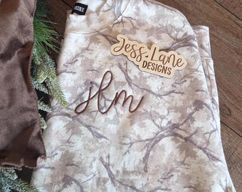 May include: A folded, long-sleeved sweatshirt with a beige and brown tree branch pattern. The sweatshirt has the embroidered initials "Hm" and a wooden tag that reads "Jess Lane Designs". A brown ribbon and greenery are in the background.