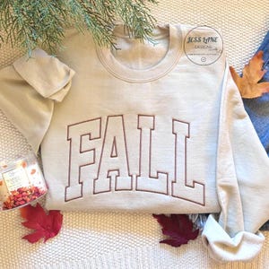 Puede incluir: Una sudadera beige de cuello redondo con la palabra FALL bordada en letras grandes de color marr&oacute;n estilo bloque en el pecho. La prenda incluye un logotipo circular en la esquina superior derecha con el texto JESS LANE DESIGNS EMBROIDERY APPAREL.