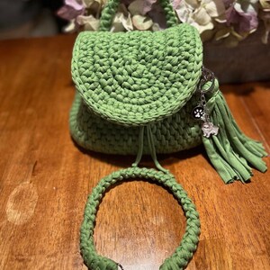 Op de afbeelding: Een groene gehaakte mini-rugzak met een lange groene gehaakte band en een zilveren bedel. De rugzak heeft een trekkoord sluiting en een klein vakje aan de voorkant.