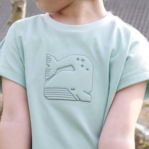 Può includere: Una t-shirt a maniche corte color verde menta chiaro con il contorno di una balena ricamato in verde scuro. Il disegno della balena è stilizzato con linee geometriche e dettagli circolari, posizionato centralmente sul petto.
