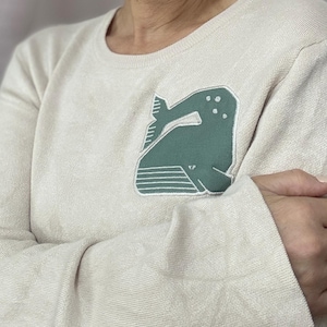 Może przedstawiać: Kremowa koszulka z długim rękawem z zieloną grafiką wieloryba na piersi. Projekt wieloryba jest obrysowany na biało i przedstawia stylizowaną ilustrację ciała i ogona wieloryba. Koszulka ma okrągły dekolt.