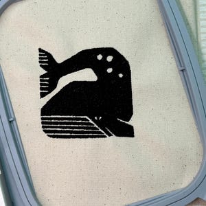 Puede incluir: Un diseño de ballena bordado en negro sobre un fondo de tela blanco. La ballena está estilizada con una forma geométrica simple y tiene algunos círculos pequeños en su espalda.