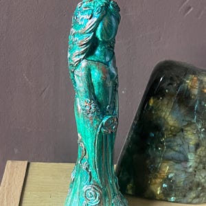 Puede incluir: Una estatua de color turquesa de una mujer con un vestido fluido, adornada con detalles florales. La estatua tiene una apariencia envejecida y antigua, con detalles en bronce. La figura est&aacute; junto a una gran piedra iridiscente.